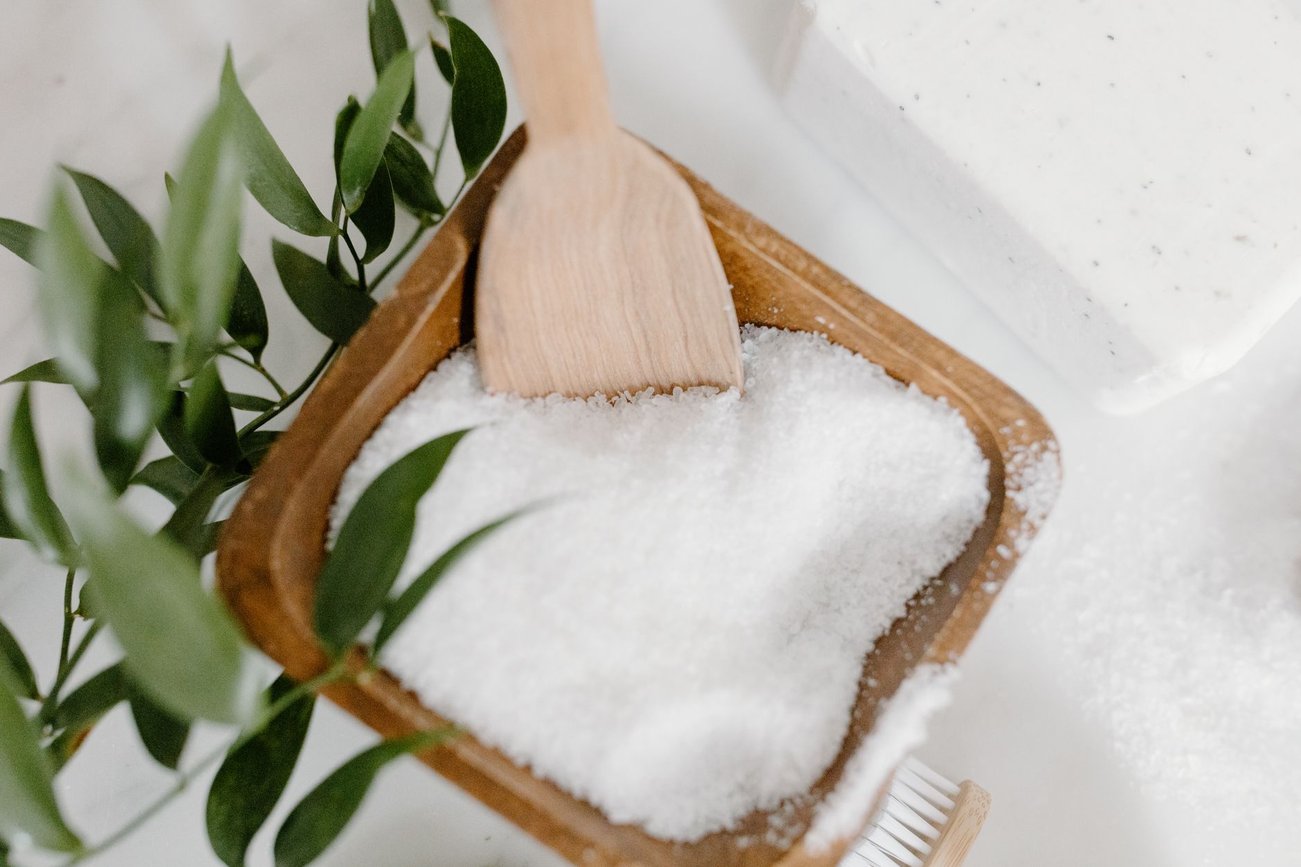 Coarse white bath salts in a wooden bowl beside a body brush and green leaves.