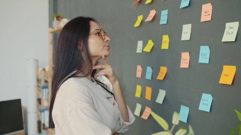 Thoughtful woman planning ideas on a chalkboard wall with colorful sticky notes in an office setting.