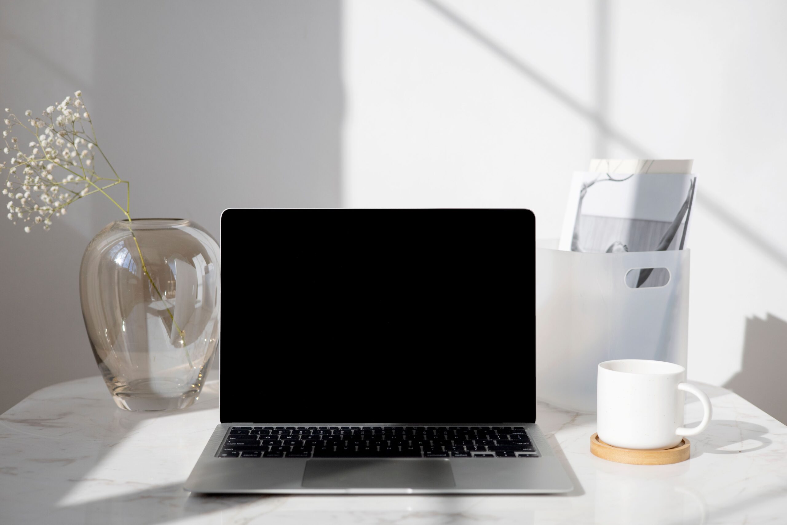 A clean modern desk with a laptop, coffee cup, vase and documents in bright daylight.
