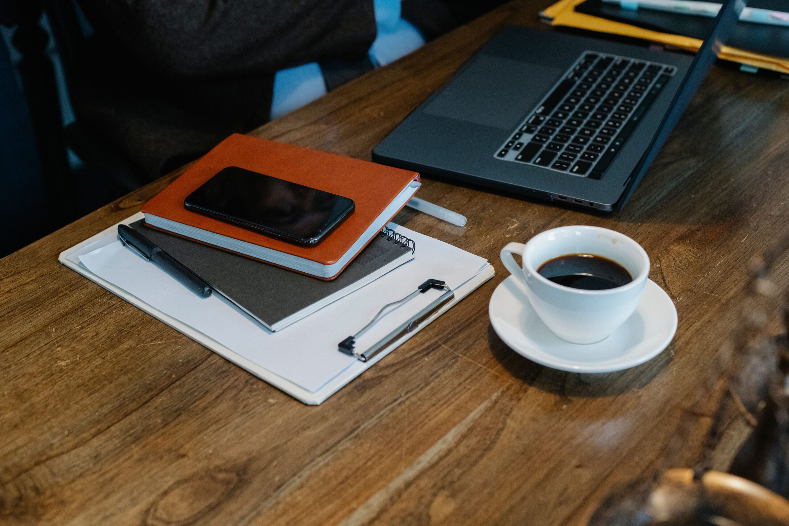 An overhead view of an open laptop, coffee, notebooks and a smartphone on a wooden desk.