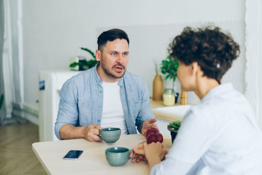 A couple sitting at a kitchen table with coffee cups, having a serious conversation.