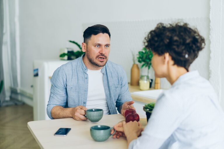 A couple sitting at a kitchen table with coffee cups, having a serious conversation.