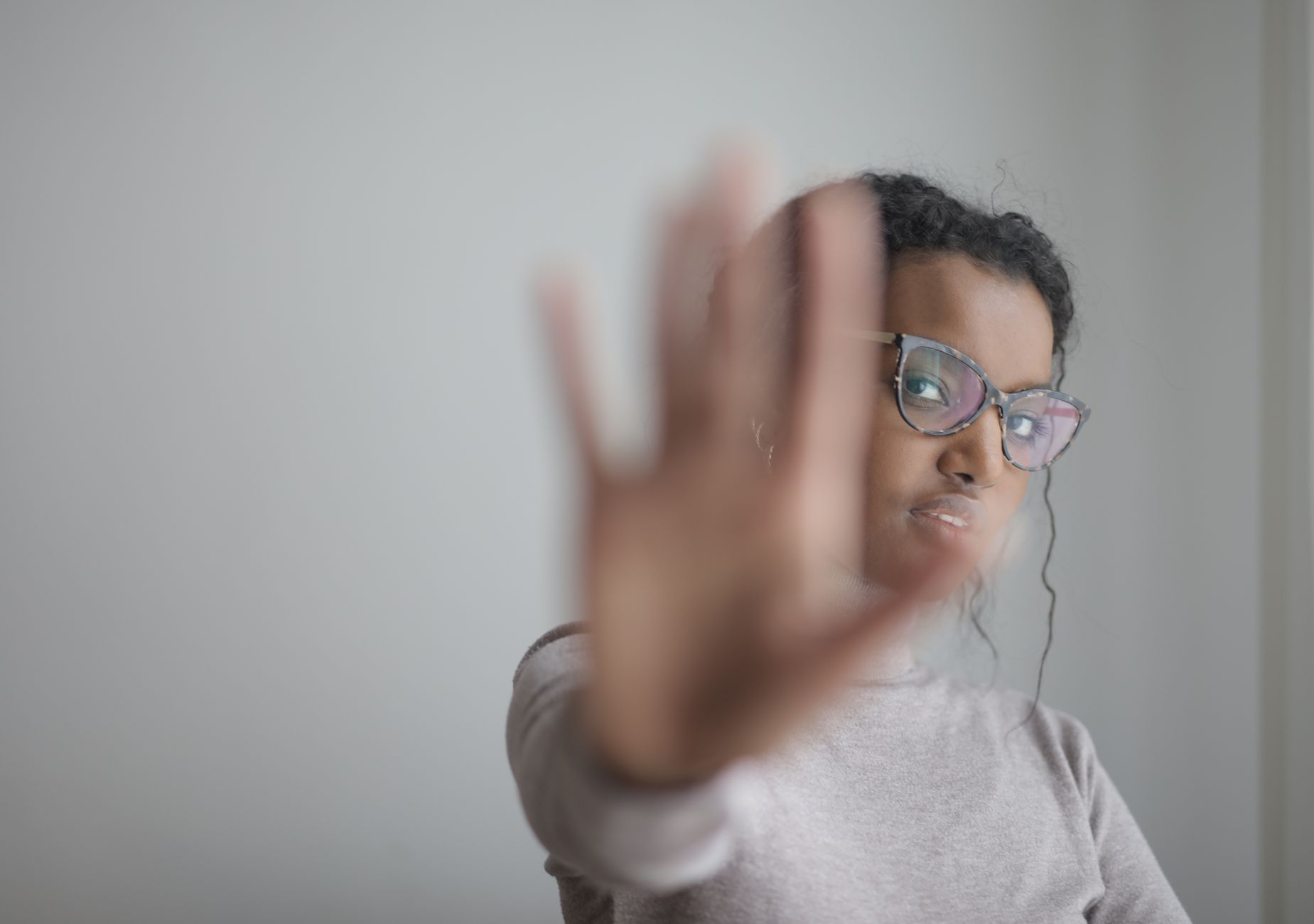 Woman in glasses calmly extending her arm in a stop gesture against a grey wall.