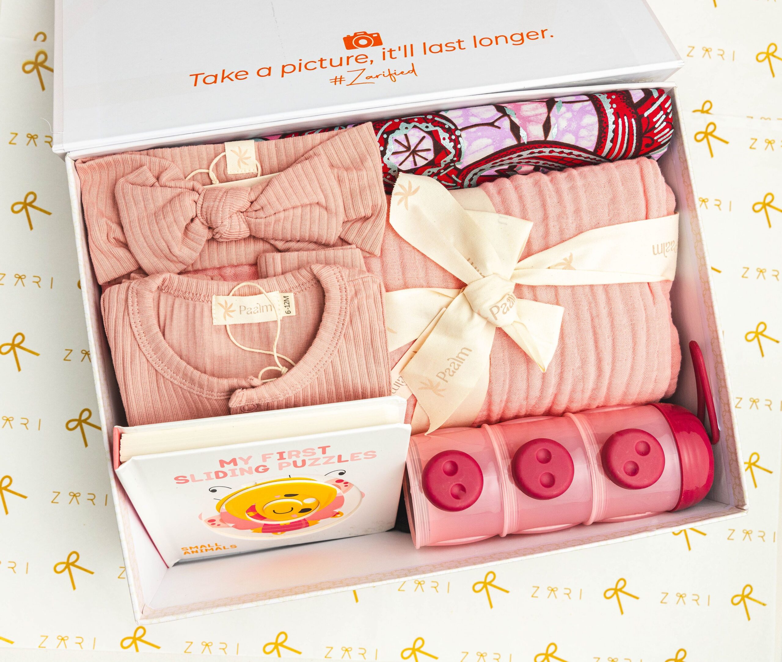 A baby gift box packed with pink clothing, a small book, and storage containers.