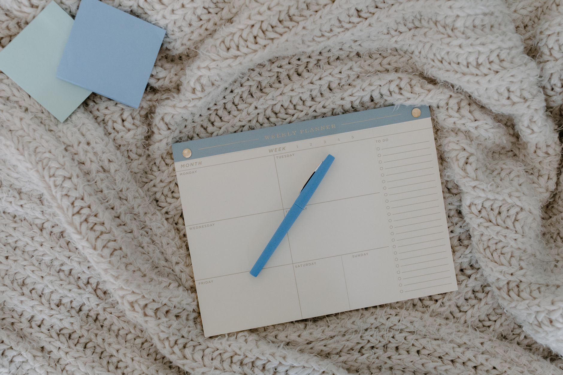A weekly planner pad and a blue pen resting on a knitted blanket beside two sticky notes.