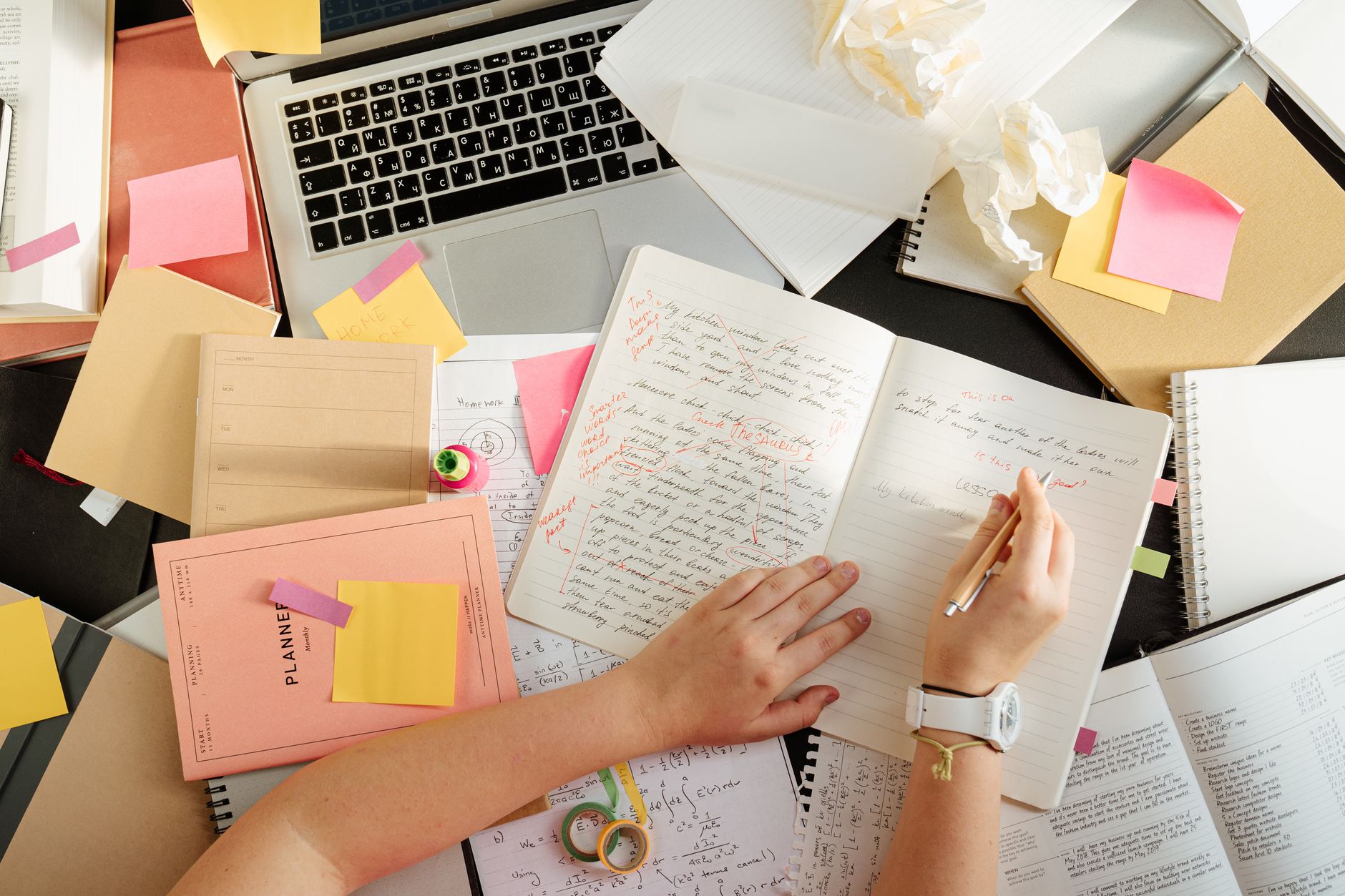 An overhead view of a cluttered desk covered with open notebooks, sticky notes, a laptop and loose papers.