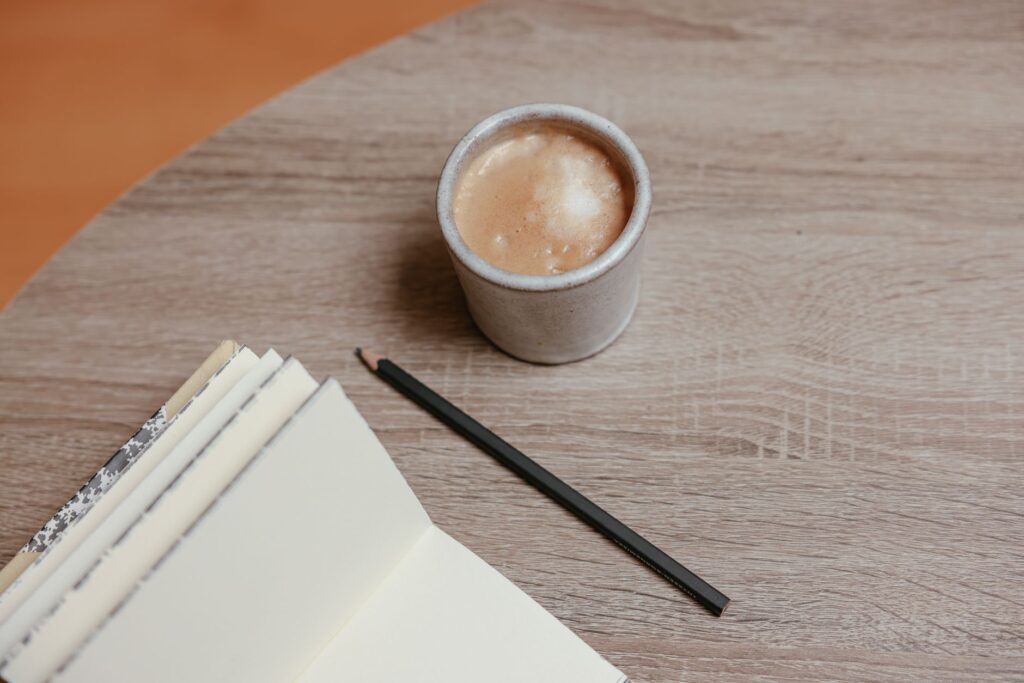 A cup of coffee, an open notebook and a sharpened pencil on a wooden table, seen from above.