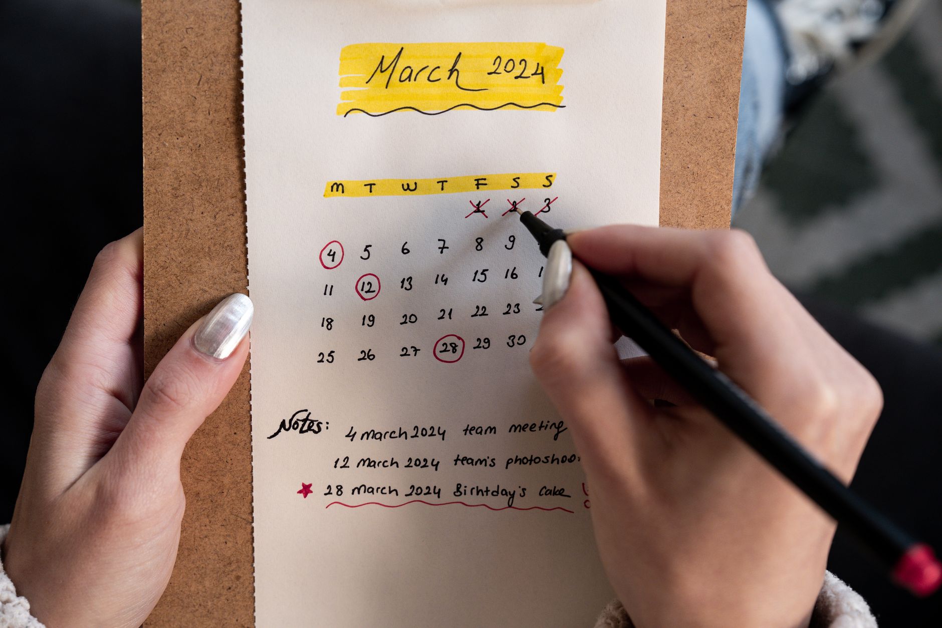 Two hands hold a clipboard with a paper calendar for March 2024, several dates circled in red.